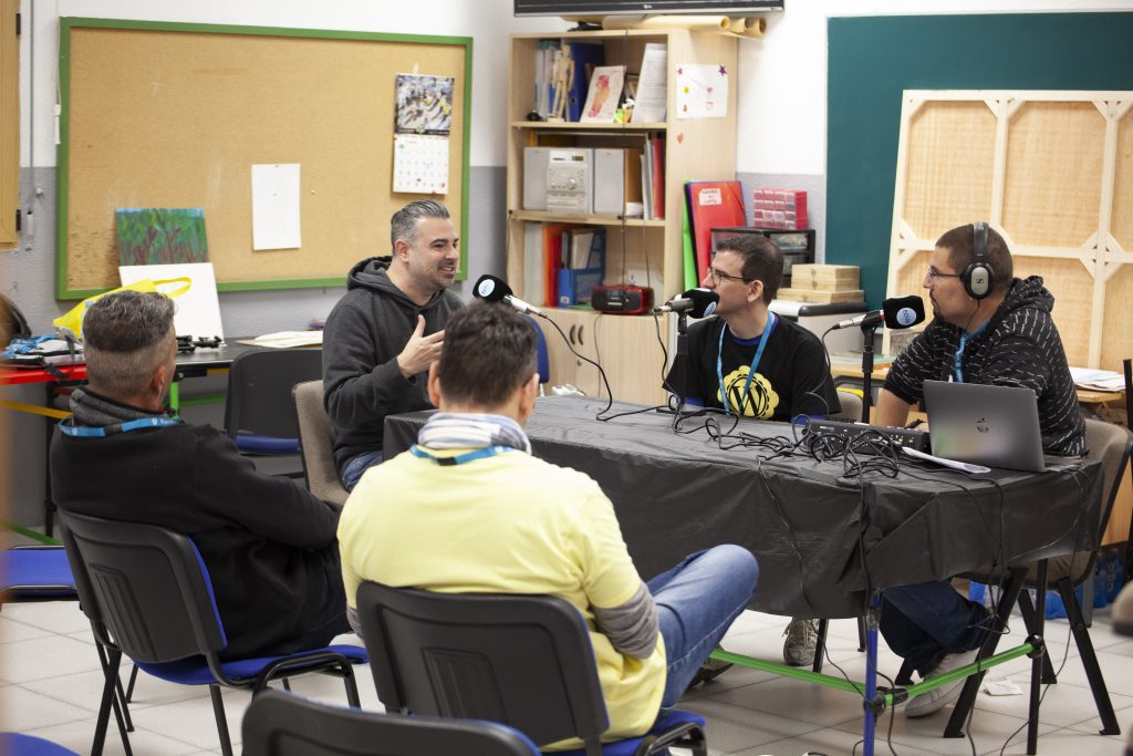 Fotografía de la sala de podcast en WordCamp Griñón con Javier Casares, David Plaza y Jorge Marín sentados a una mesa, con tres micrófonos, grabando el podcast y con varios asistentes sentados escuchando.